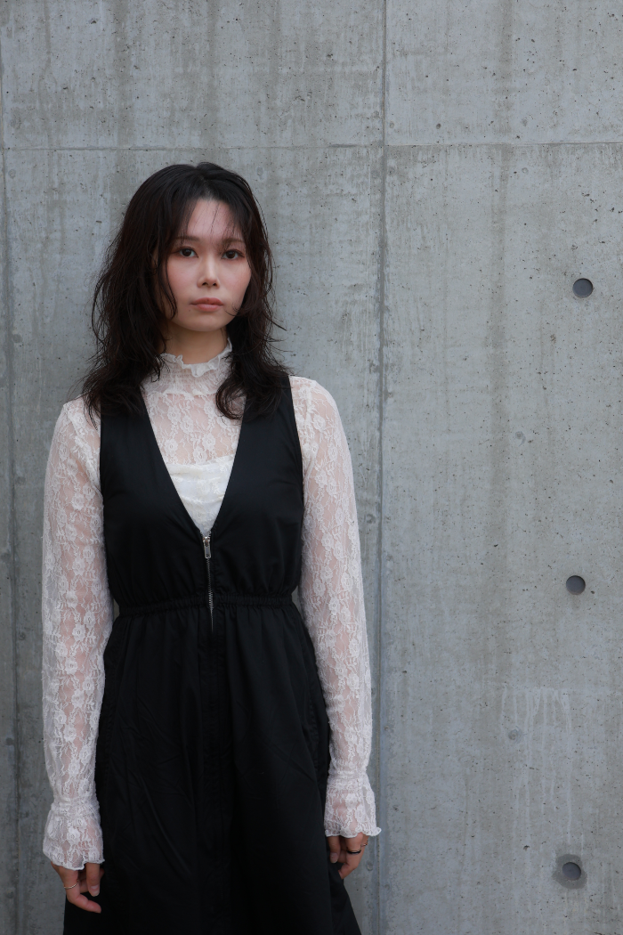 札幌市東区の美容室「ハートシェイプドボックス」のリケラトリートメント紹介イメージ。コンクリート壁を背景に、黒いベストと白いレースブラウスを着た女性が凛とした表情でカメラを見つめる。リケラで艶やかに整えられたダークブラウンの髪が、大人の女性の本質的な輝きを表現。