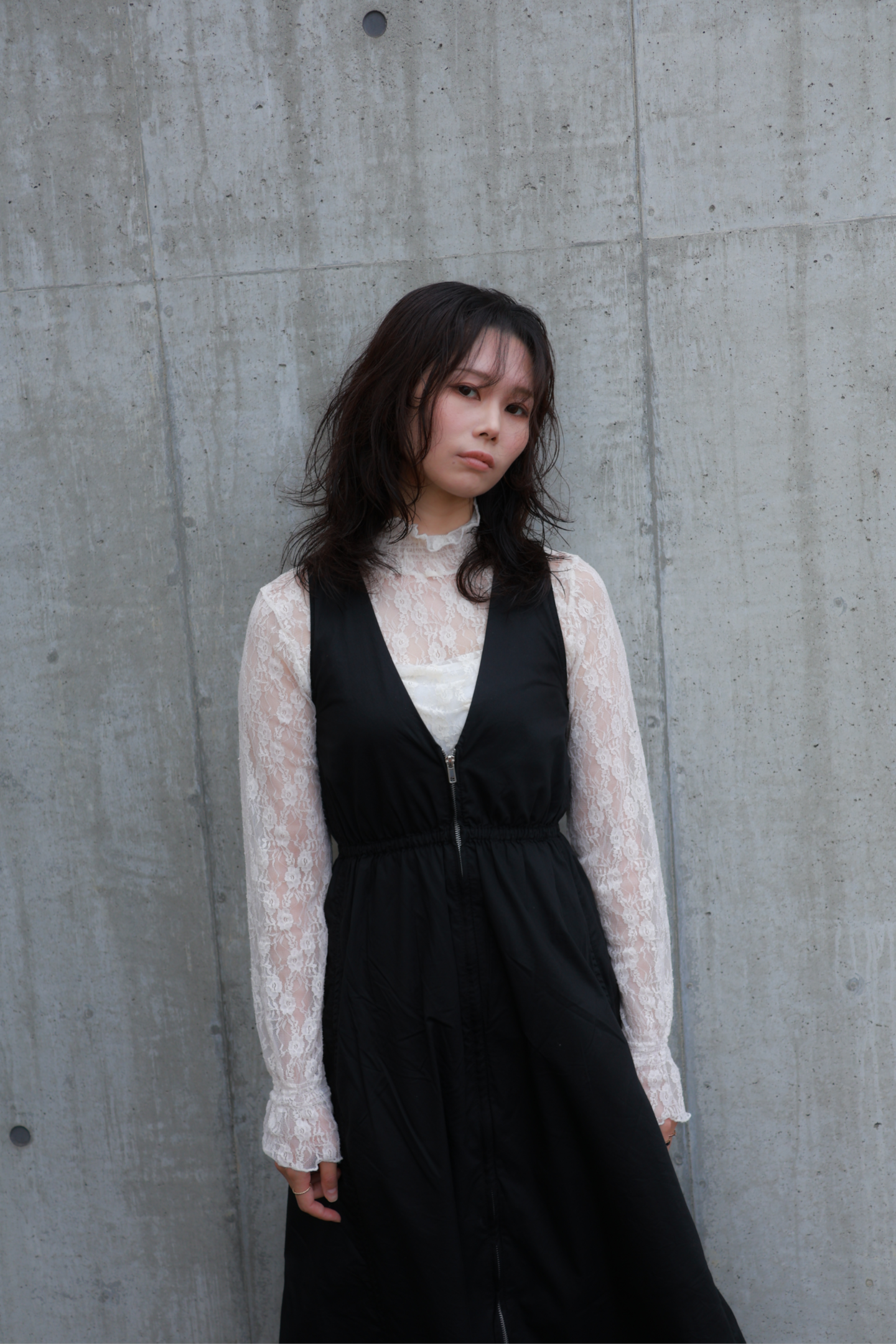 札幌市東区の美容室「ハートシェイプドボックス」のリケラトリートメント紹介イメージ。コンクリート壁を背景に、黒いベストと白いレースブラウスを着た女性が凛とした表情でカメラを見つめる。リケラで艶やかに整えられたダークブラウンの髪が、大人の女性の本質的な輝きを表現。