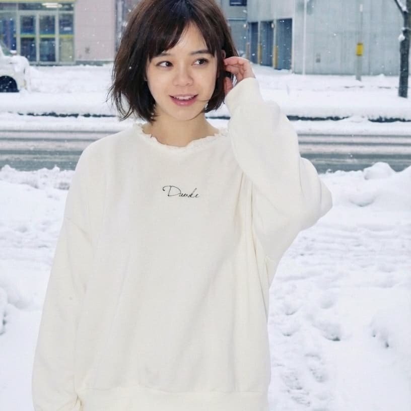 札幌市東区の美容室「ハートシェイプドボックス」のヘアスタイル作例。雪景色が広がる札幌の街角で、クリーム色のニットとチェックスカートを着て明るい笑顔を見せる女性。冬のファッションに似合うナチュラルなヘアスタイル。